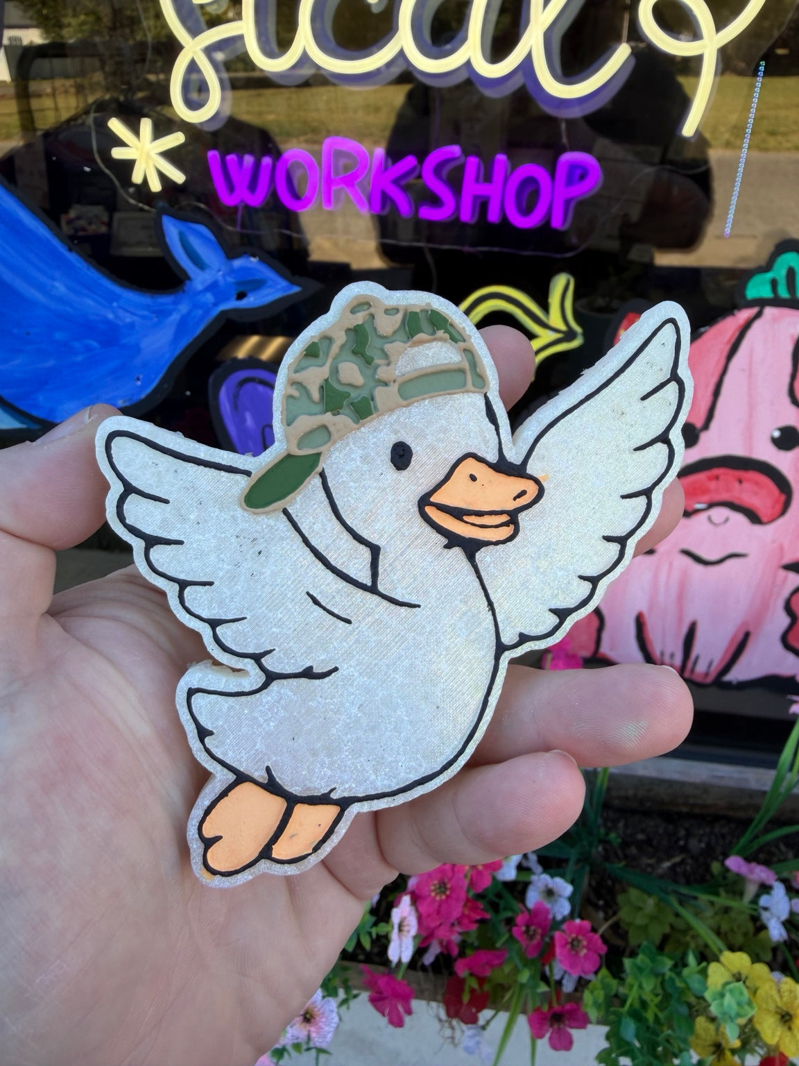 Handmade duck in camo hat freshie with wings outstretched, photographed in front of Whimsical Workshop