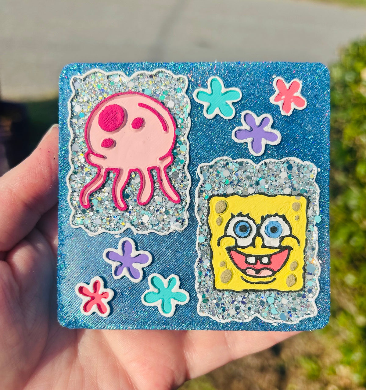 Hand-painted SpongeBob and pink jellyfish car freshie with blue glitter background, sparkly panels and turquoise, purple and pink sea flowers